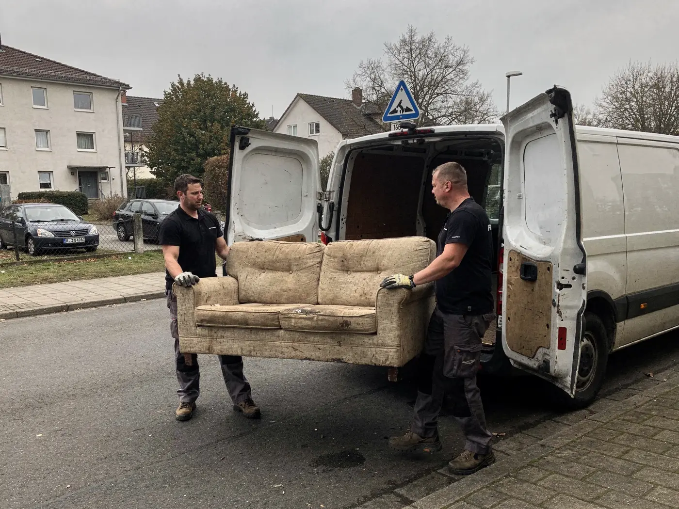 Zwei Mitarbeiter tragen ein altes Sofa zu den offenen Hecktüren des Transporters.